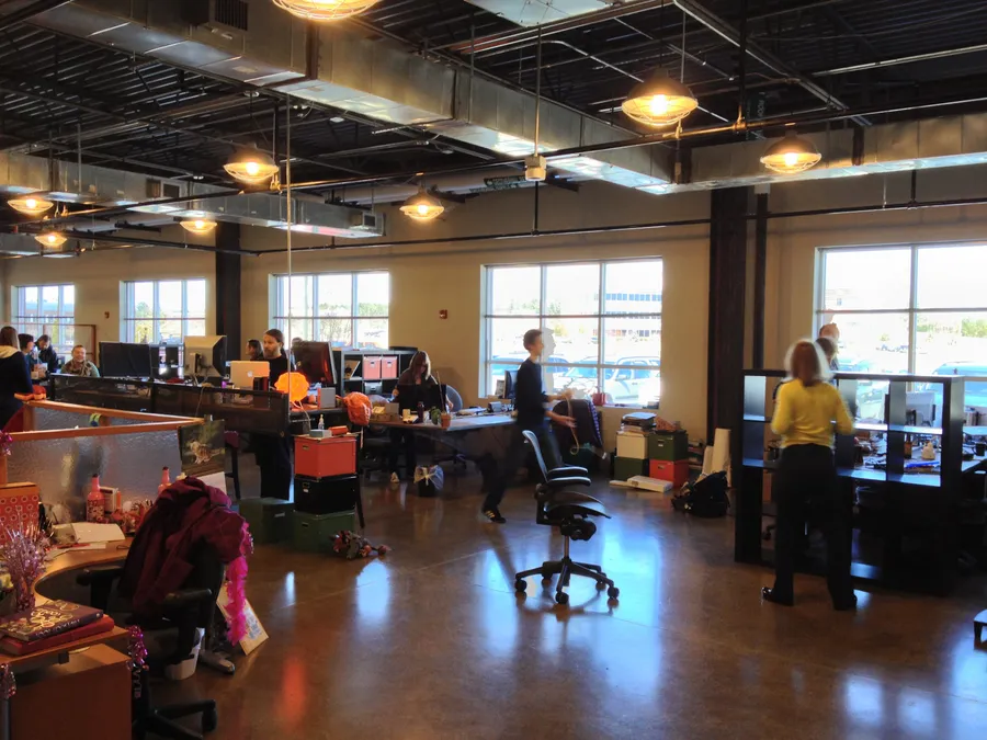 Cavernous office space with long desks, concrete floors, and exposed pipes in the ceiling