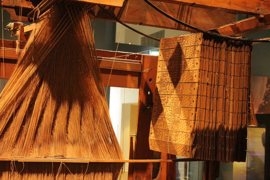 A closeup photo of a Jacquard loom, a large wooden contraption with many threads attached and a thick stack of punch cards.