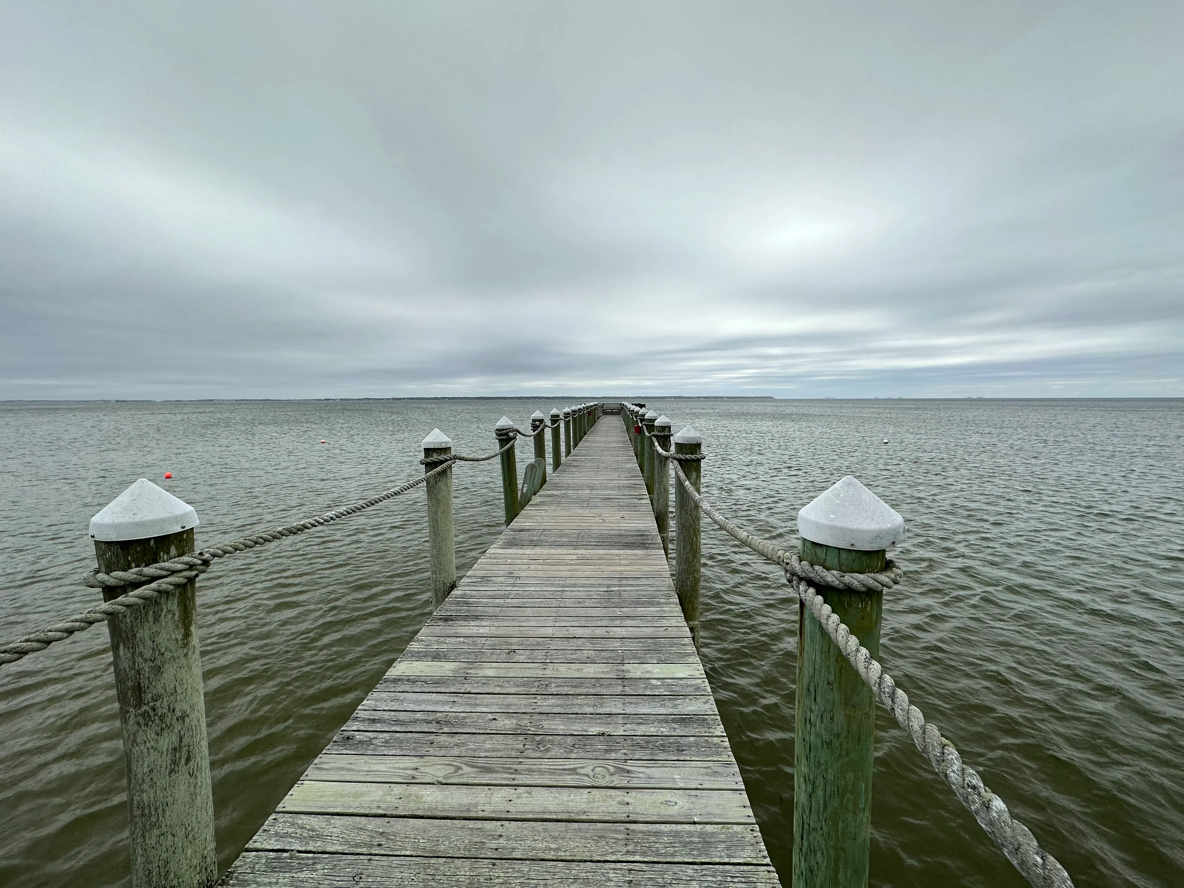 A weathered pier stretches out across a calm water in an overcast sky.