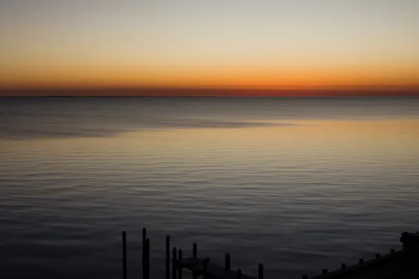 Sunset over a large, mostly still body of water. The sun is already below the horzion, so the water is dark and the sky is fairly dark as well. There are a few wisps of clouds near the horizon that lighten the burt orange of the sky there.